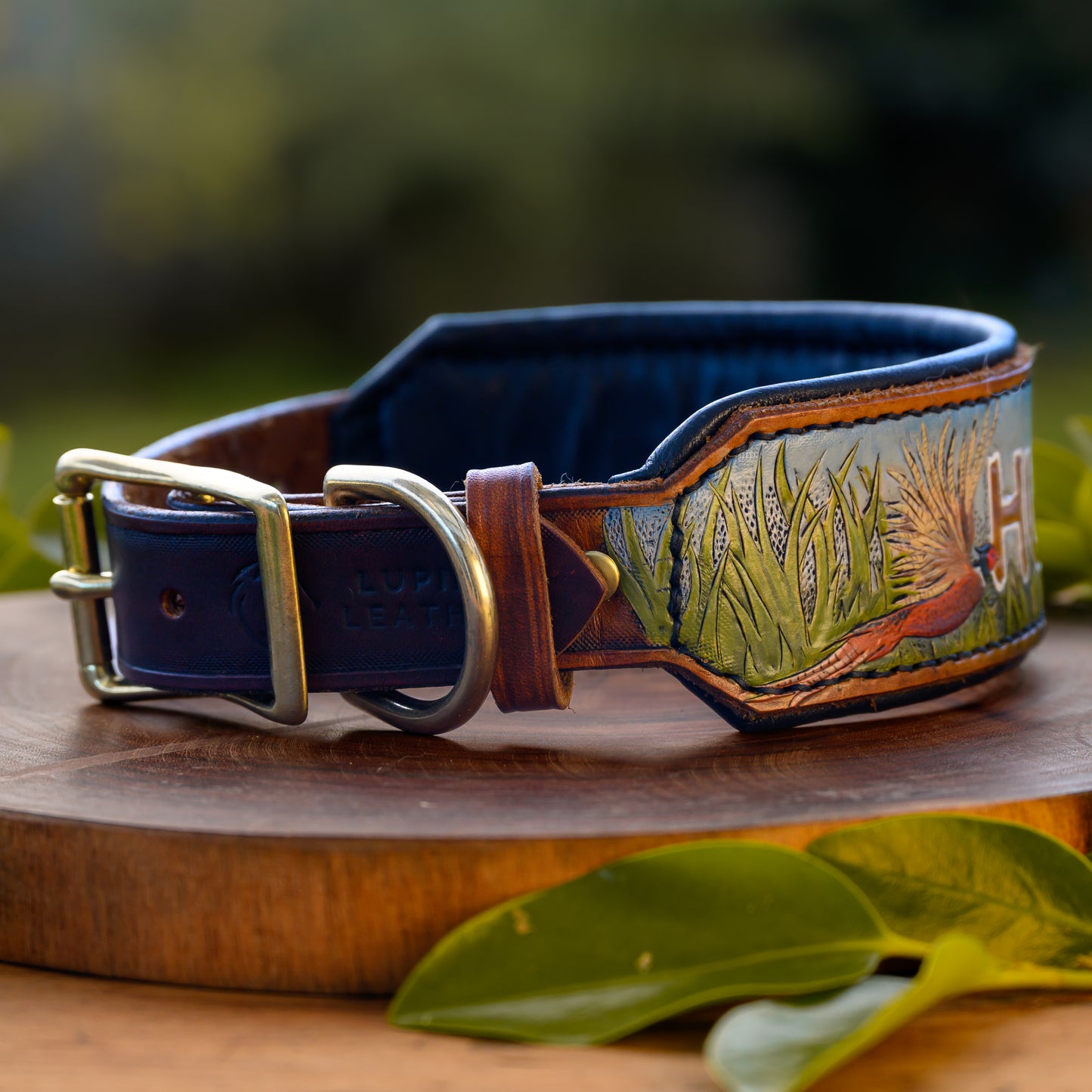 A custom hand-tooled leather dog collar with a black interior, brass buckle, and detailed carving of a pheasant and grass, displayed on a wooden block with green leaves.
