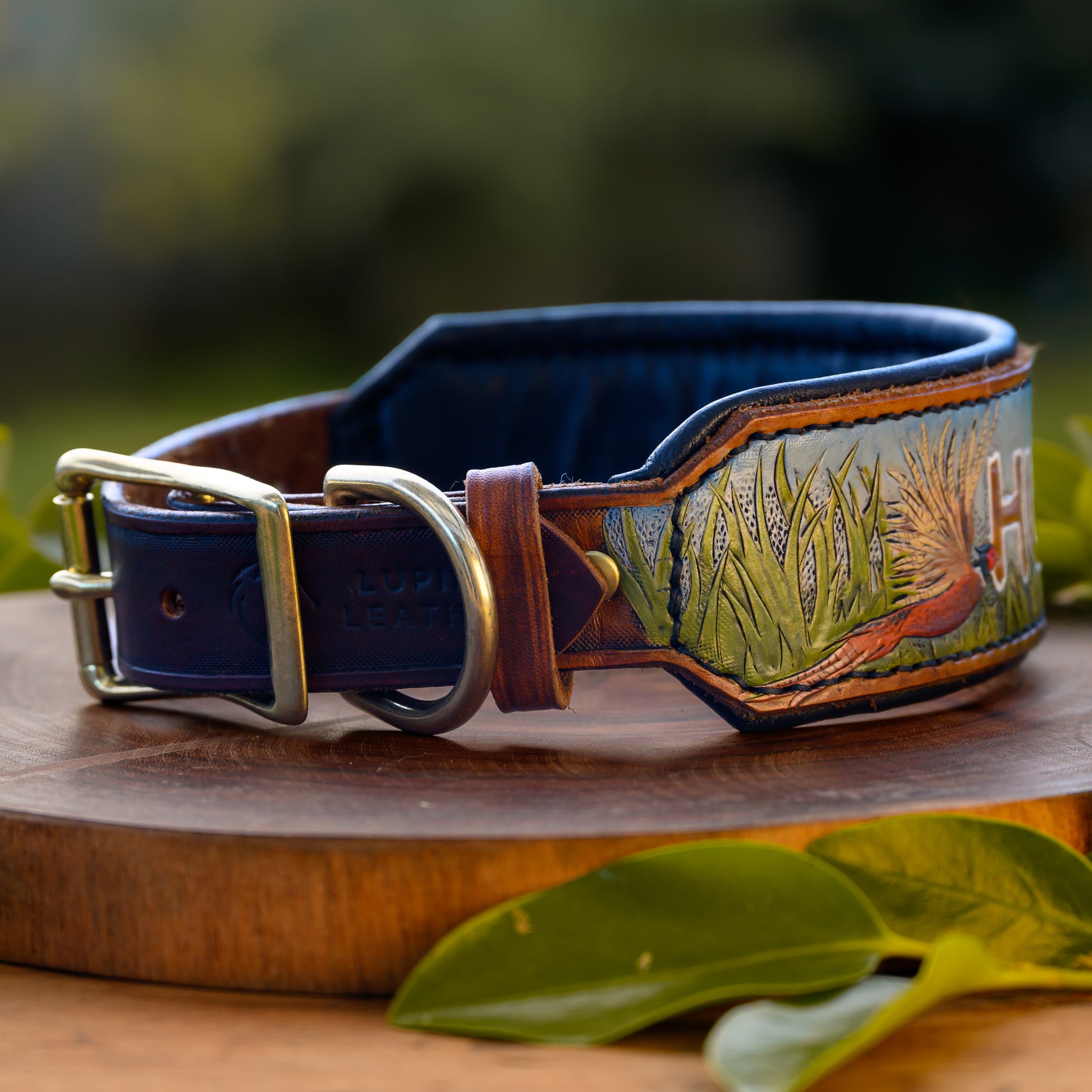 A custom hand-tooled leather dog collar with a black interior, brass buckle, and detailed carving of a pheasant and grass, displayed on a wooden block with green leaves.