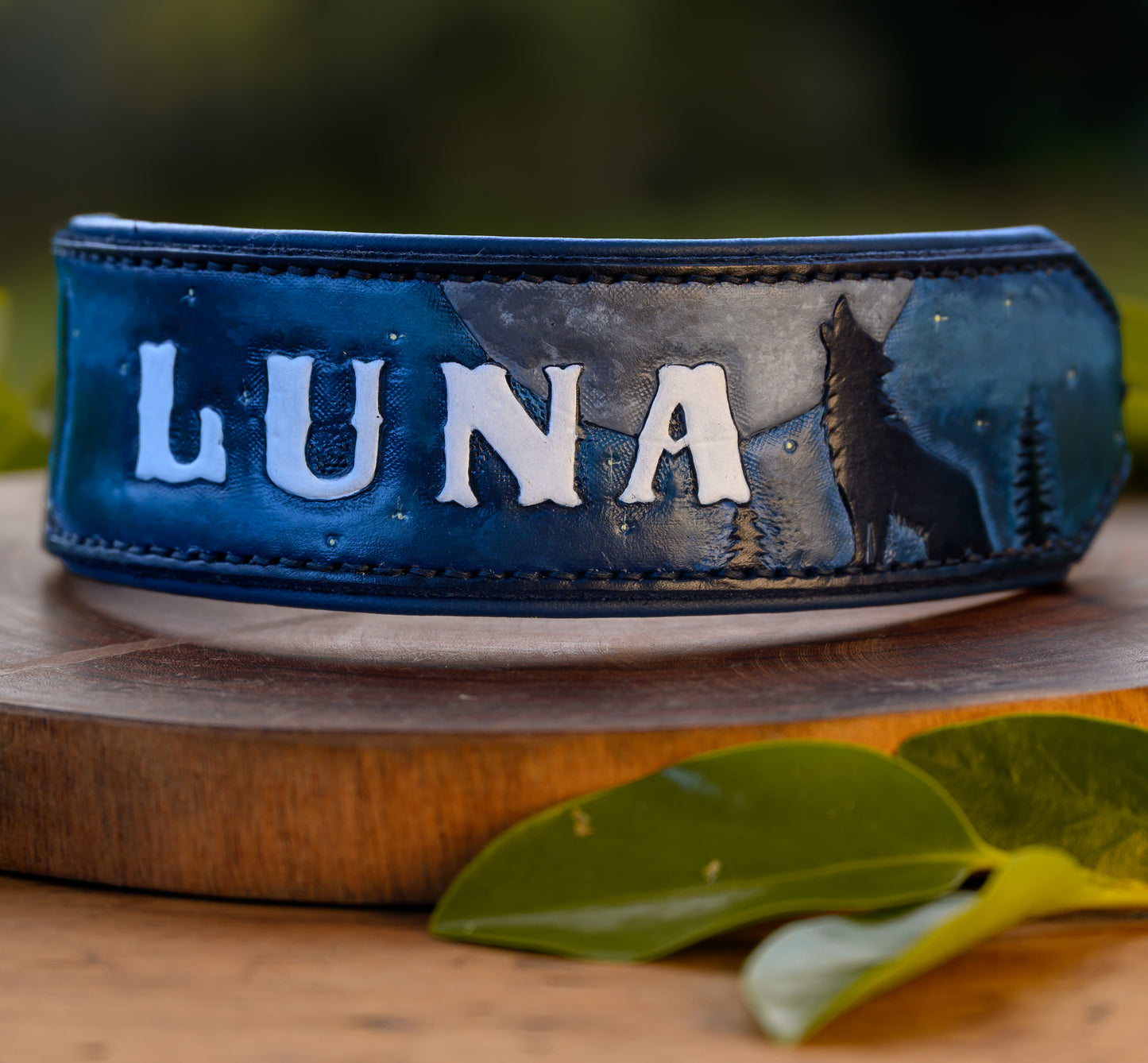 A blue leather collar with 'LUNA' in raised white letters, featuring a dark blue and gray wolf silhouette design, displayed on a wooden stand with green leaves in the foreground.