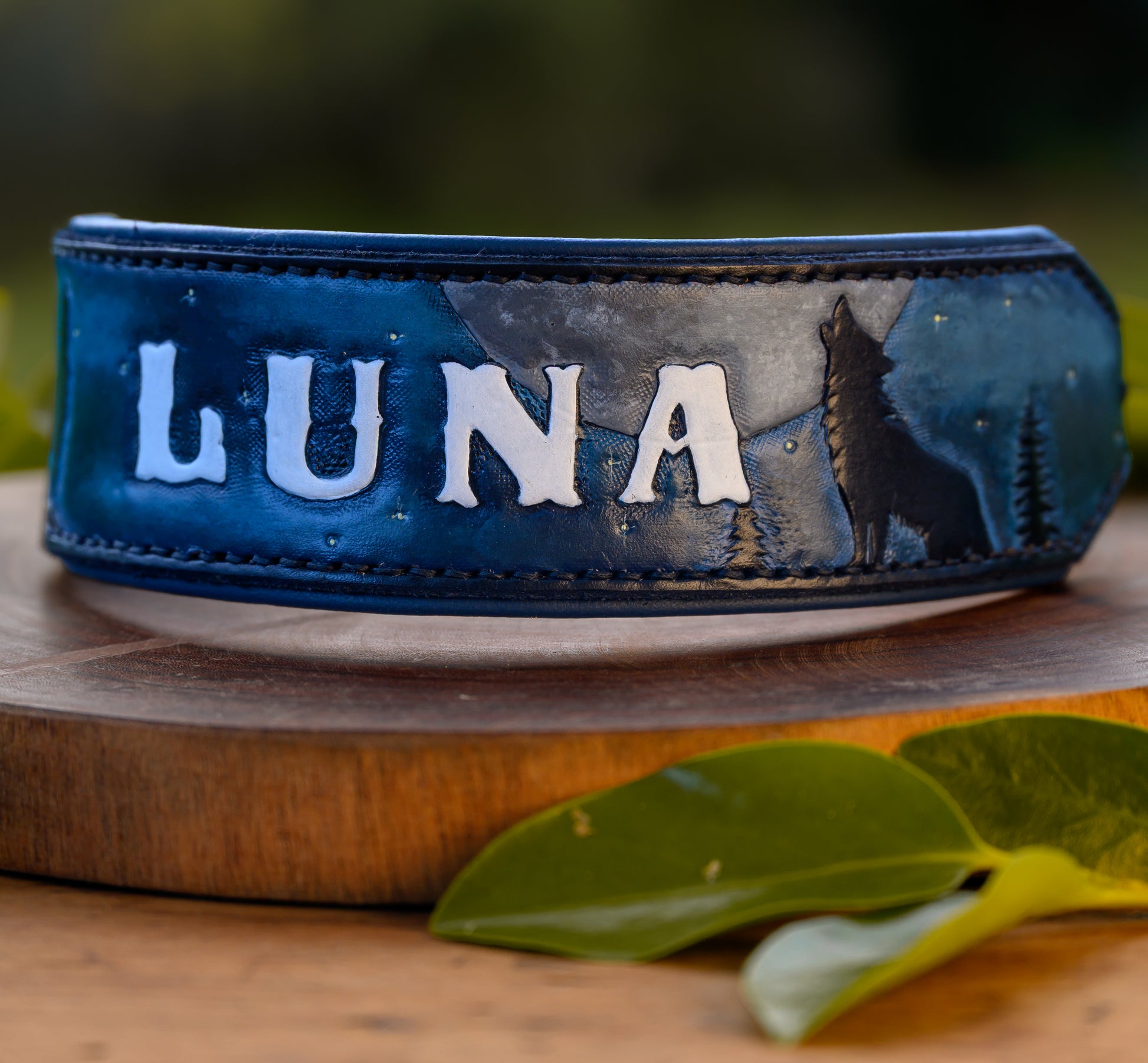 A blue leather collar with 'LUNA' in raised white letters, featuring a dark blue and gray wolf silhouette design, displayed on a wooden stand with green leaves in the foreground.